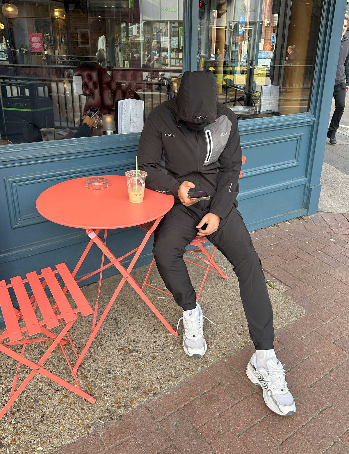 Person sitting at a red table outside a cafe, wearing a black hoodie and pants.
