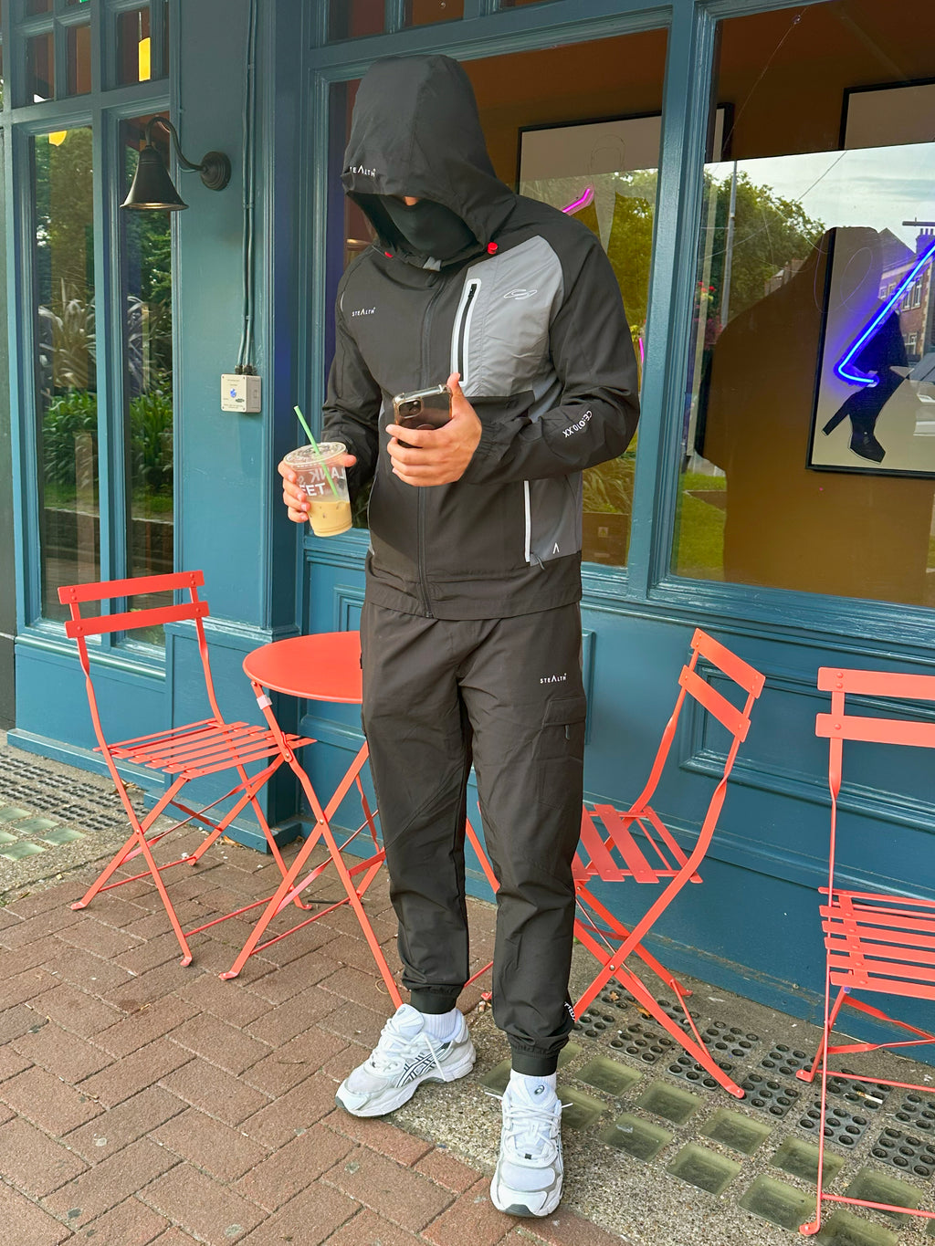 Person in black hoodie and pants standing outside a building with red chairs and a blue wall.